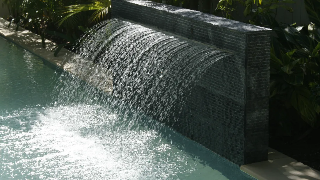Honed black stone water blade spilling a clear sheet of water into a pool edged with tropical planting